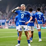 Soccer player in a blue kit with number 9 poses with a hand to the ear as teammates celebrate on the field behind him.