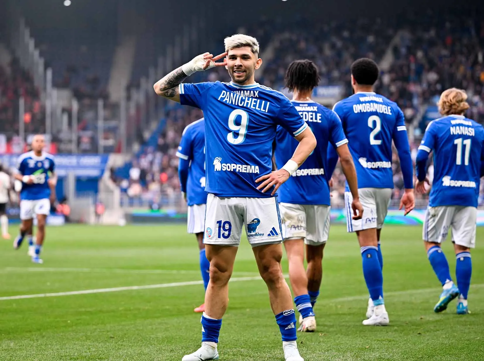 Soccer player in a blue kit with number 9 poses with a hand to the ear as teammates celebrate on the field behind him.