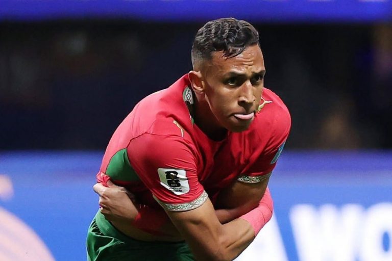 Soccer player in red jersey and green shorts bending forward, balancing on one leg during a match on a green field.",