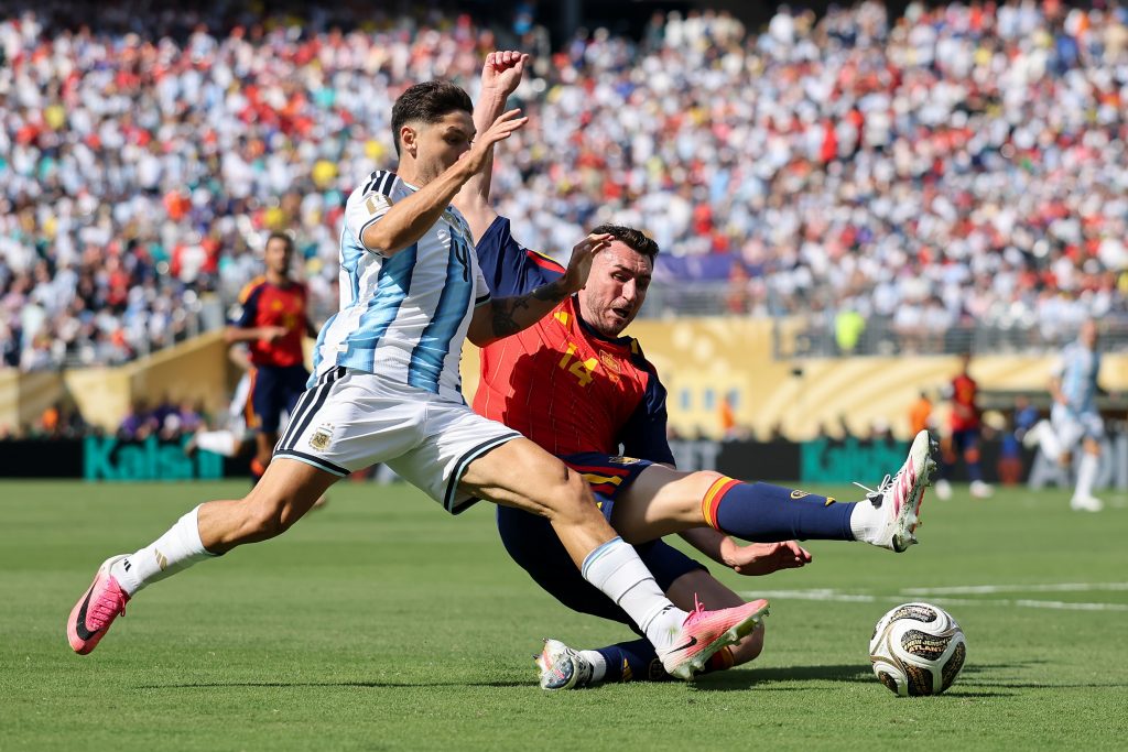 Two soccer players compete for the ball during a match; one wears a light-blue/white Argentina kit and the other a red/blue jersey, with a full stadium in the background.